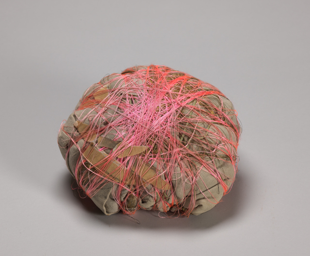 A small parcel covered in taupe cloth and wrapped in criss-crossing bright pink synthetic thread. The parcel is photographed against a grayish-white background and casts a shadow.