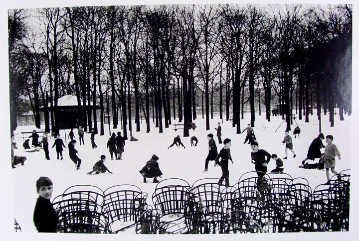 A snowy park scene with several children playing in the snow. In the foreground, there are stacked metal chairs. The background includes bare trees, and children are engaged in activities such as sledding and playing. The scene is primarily white with dark silhouettes of the trees and children.
