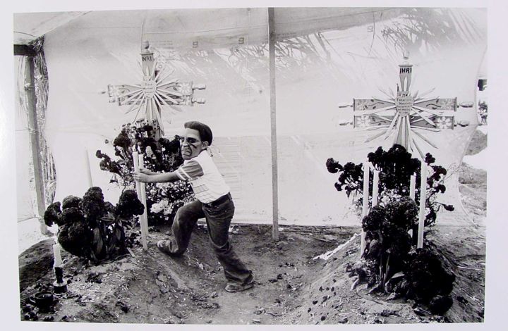 A figure wearing glasses and a striped shirt stands between two large crosses in a dirt area. The crosses are decorated and surrounded by dark flowers. In the background, there is a white tent-like structure, and candles are placed near the crosses.