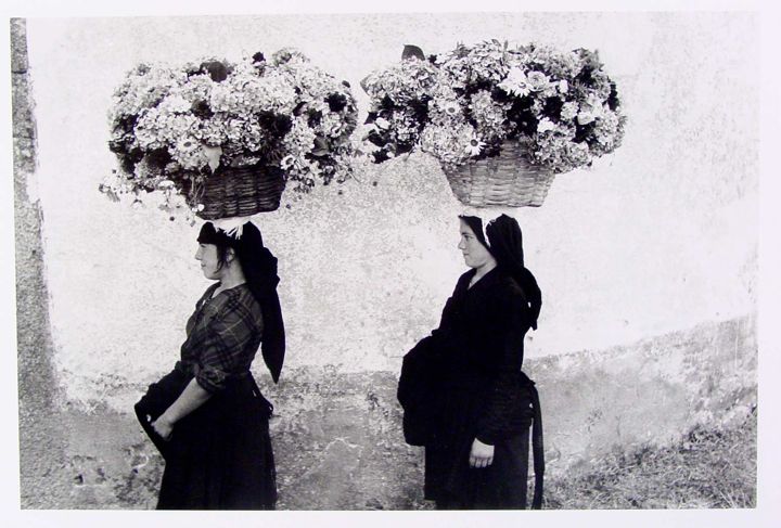 Two women stand side by side in front of a textured wall. Each woman has a large woven basket filled with flowers on her head. One woman wears a checkered top, while the other is dressed in a long black dress.