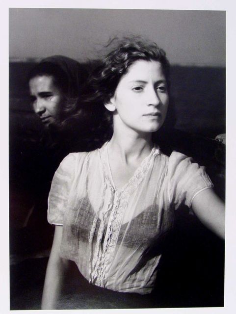 Two women are depicted in the image. The woman in the foreground has light-colored hair and is wearing a light, sheer blouse. She is looking slightly to the side, with her hair being moved by the wind. In the background, there is a blurred landscape, likely water, with a darker figure of the second woman partially visible.