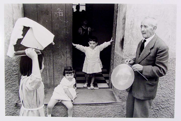 A black-and-white image shows a man in a suit holding a balloon, standing on a stone surface. To the left, a girl with a paper hat is present, while another girl in a light dress sits on the ground. A third child stands in a doorway, extending their arms. The background includes a wooden door and a checkered floor visible inside the building.