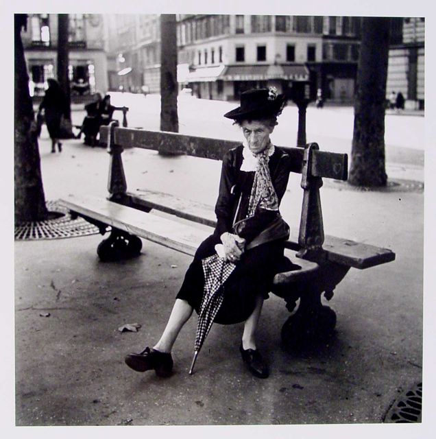 A black and white photograph depicting an elderly woman seated on a wooden bench in a public area. She is dressed in a dark outfit and a large hat, holding a closed umbrella. The background includes trees and buildings, with a few distant figures. The ground is paved, covered with scattered fallen leaves.