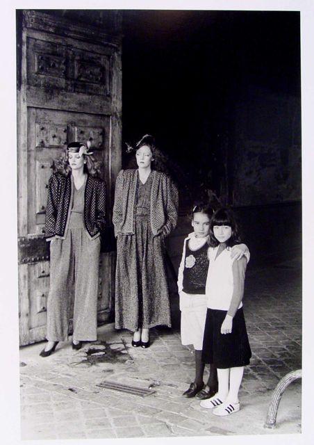 Two adult women and two young girls stand in front of a large wooden door. The women wear patterned outfits, while the girls are dressed in a mix of light and dark clothing. The scene is dimly lit, with a tiled floor visible beneath them. The door and clothing display various textures.